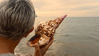 Eine ältere Dame lauscht am Meer an einer Muschel