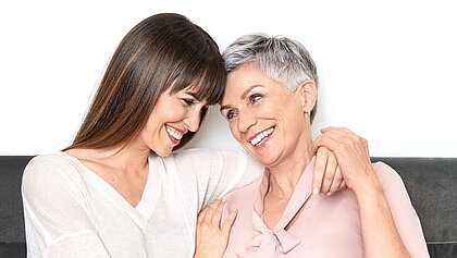 Eine ältere und junge Dame sitzen gemeinsam auf einem Sofa und halten sich lächelnd im Arm